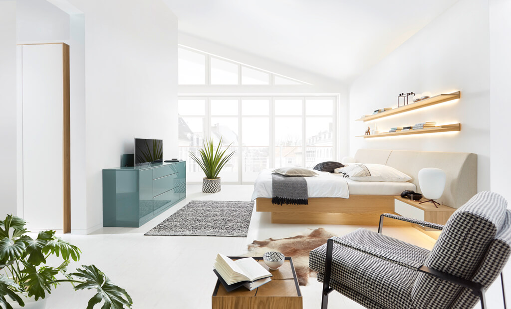 Schlafzimmer komplett in weiß mit grün/blauem Sideboard mit Fernseher, der gerade aus dem Sideboard heraus fährt.